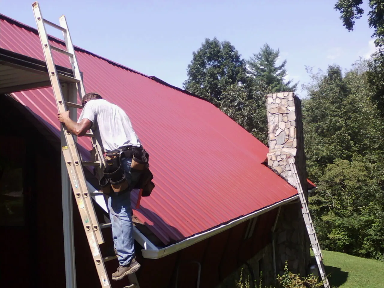 Roofer on a ladder beside a red metal roof and stone chimney — Soffit & Fascia Repair by Roofing Brothers in Red Creek